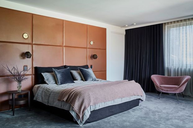 A sophisticated bedroom with a large upholstered bed dressed in layered grey and mauve bedding, set against a dramatic copper-toned padded feature wall with integrated wall lights. Floor-to-ceiling dark curtains frame sheer window coverings, while a sculptural mauve lounge chair and textured grey carpet complete the moody, refined interior.