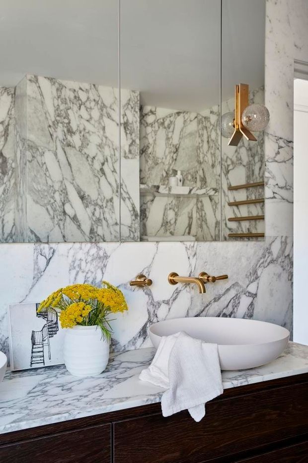 A refined bathroom vanity with extensive white marble surfaces featuring dramatic grey veining. A white vessel basin sits on a marble benchtop above dark timber cabinetry, paired with brushed brass wall-mounted tapware. A large mirrored cabinet reflects marble shelving, while a sculptural brass-and-glass wall light adds a luxe accent.