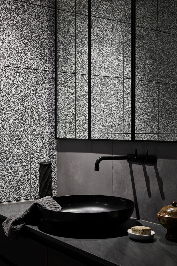 A dramatic, moody bathroom vanity scene featuring a large matte-black vessel basin on a dark stone benchtop, a black wall-mounted tap, and dark cabinetry. The walls combine grey concrete-look tiles with striking terrazzo-style patterned panels framed by black metal trims. Strong directional light creates pronounced shadows, while minimal accessories add a refined, monochromatic feel.