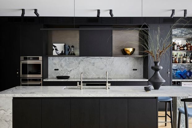 A sophisticated contemporary kitchen dominated by a large white marble island and matching marble splashback. Matte dark cabinetry, integrated stainless-steel appliances, open shelving, and sculptural black pendant-free styling create a dramatic, refined look. Bar stools and a drinks display are visible at the right.