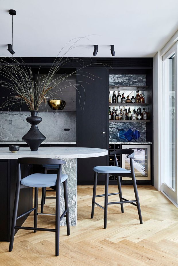 A sophisticated open-plan home bar and kitchen entertaining area with a round marble-topped island, dark cabinetry, integrated backlit liquor shelving, and sculptural black pendant and track lighting. Pale herringbone timber flooring contrasts with the black joinery and veined stone surfaces, while bar stools and a large decorative vase complete the refined interior.