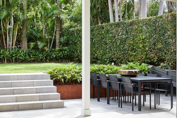 A landscaped outdoor entertaining area with a tiled patio, dark outdoor dining table and chairs, raised planter beds, and broad steps leading into a lush tropical garden. A white structural column divides the patio from the garden view, while dense hedging and palms create a private setting.