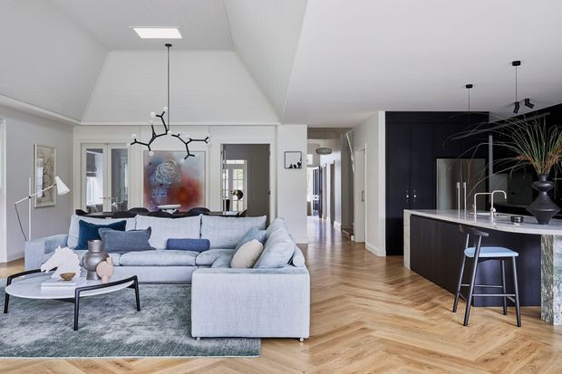 A bright, open-plan contemporary living and kitchen area with white walls and ceilings, a large pale sectional sofa, patterned rug, sculptural pendant lighting, and herringbone timber flooring. The kitchen features dark cabinetry, a light stone island with integrated sink, and bar seating. Glazed doors and a hallway extend into the background.