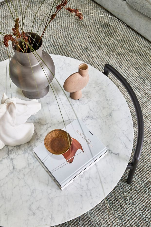 Close-up overhead view of a round white marble coffee table with a black metal edge and legs, styled with sculptural ceramic vessels, a metallic vase containing dried grasses, a white decorative object, and an art book. The table sits on a textured grey patterned carpet beside a light grey upholstered sofa. The styling is refined and contemporary with muted neutral tones and warm metallic accents.