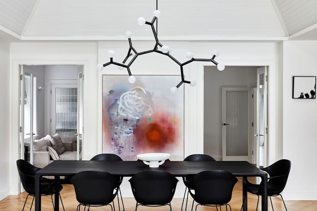 Bright contemporary dining room with a long dark dining table surrounded by black sculptural chairs. A large abstract artwork anchors the wall, while a sculptural black-and-white pendant light hangs beneath a white vaulted ceiling. Symmetrical doorways lead to adjacent rooms, with light timber flooring and white architectural detailing.