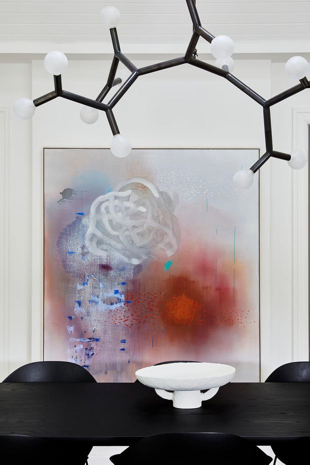 A refined dining room centered on a long black dining table with black sculptural chairs and a white sculptural bowl. A large abstract artwork in soft blue, pink, red, and white tones is mounted against white wall panelling, while a dramatic black branching pendant light with spherical white bulbs hangs overhead. The composition is contemporary and art-focused, with a restrained monochrome base and colourful artwork accents.