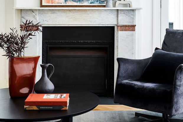 A refined living room vignette centred on a black traditional fireplace with a pale marble mantel and white wall panelling. A deep navy upholstered armchair, round black coffee table, dark ceramic vase, red planter and stacked books create a sophisticated, high-contrast setting with warm timber flooring visible beneath.