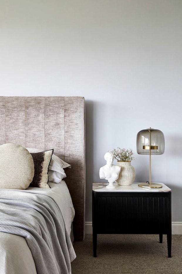 A refined bedroom vignette featuring a tall upholstered, channel-panelled headboard, layered neutral bedding, and a black fluted bedside cabinet with a pale stone-like top. A sculptural white bust, ceramic vase with dried flowers, and a smoked-glass and brass table lamp sit on the nightstand. The room has light grey walls, neutral carpeting, and a calm, softly lit contemporary aesthetic.