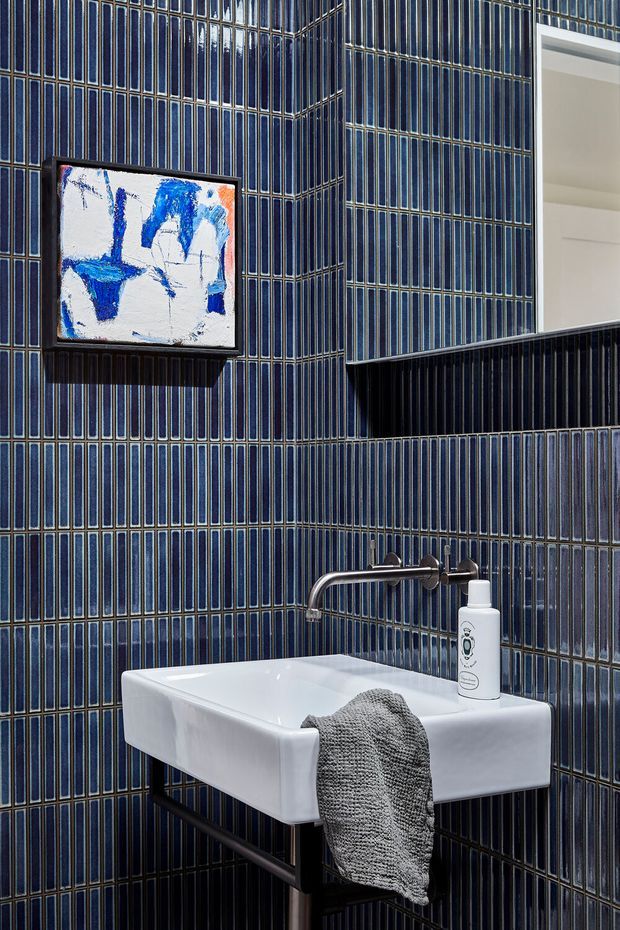 A compact, highly designed bathroom centered on a wall-mounted white rectangular basin with exposed dark metal support framing and a wall-mounted chrome tap. The walls are fully clad in narrow vertically arranged glossy dark blue tiles with contrasting light grout. A black-framed abstract artwork hangs above the basin, while a recessed black shelf and partial mirror or opening add depth. The space has a dramatic, refined, graphic appearance.