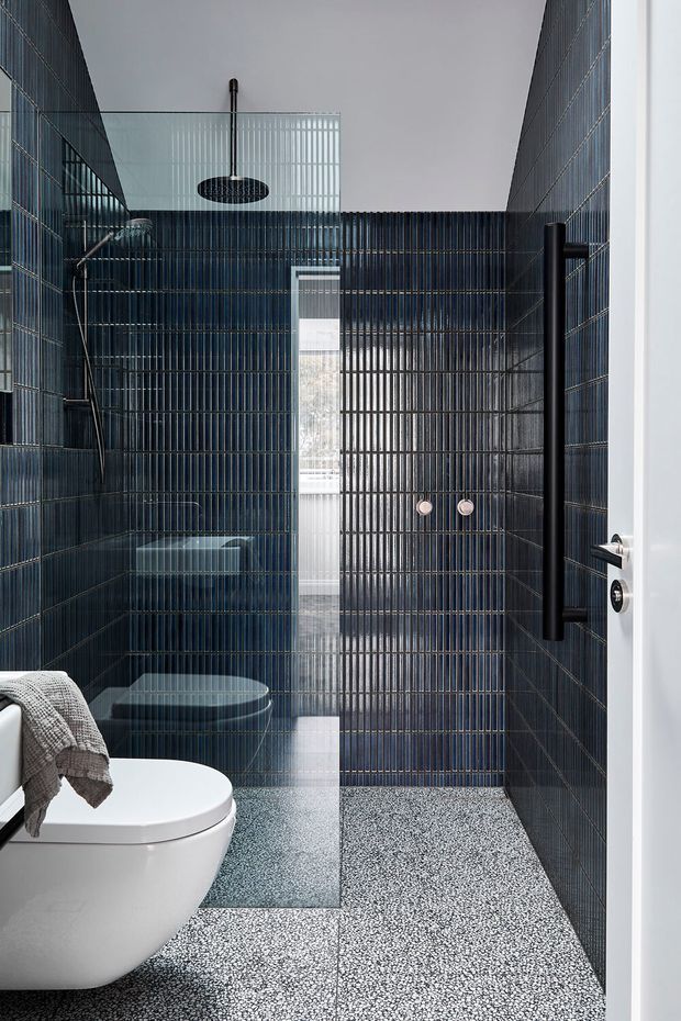 A compact contemporary bathroom viewed from the doorway, featuring dark blue vertically ribbed wall tiles, a matching tiled shower enclosure, black fixtures and fittings, a wall-mounted toilet, and a speckled terrazzo-style floor. A clear glass shower screen and white door frame add contrast, while a small high window brings in natural light.