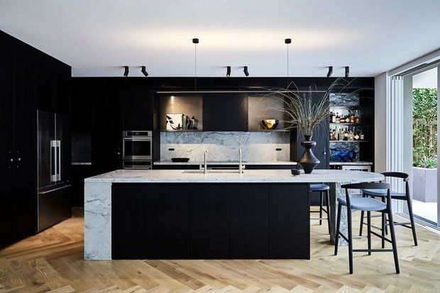 A sophisticated contemporary kitchen with a large black island, dark full-height cabinetry, integrated appliances, and a pale veined stone benchtop and splashback. The open shelving displays glassware, while bar stools sit at the island. Herringbone timber flooring, minimalist track and pendant lighting, and full-height glazing create a refined, high-contrast interior.