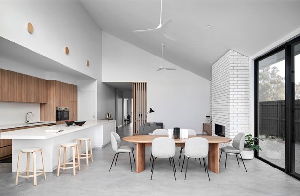 Bright open-plan contemporary living space combining a kitchen, dining area and lounge. The room features a white kitchen with warm timber cabinetry, a large island with timber stools, a timber dining table with upholstered chairs, polished concrete flooring, white walls, exposed white brick around the fireplace, ceiling fans, and expansive black-framed glazing.