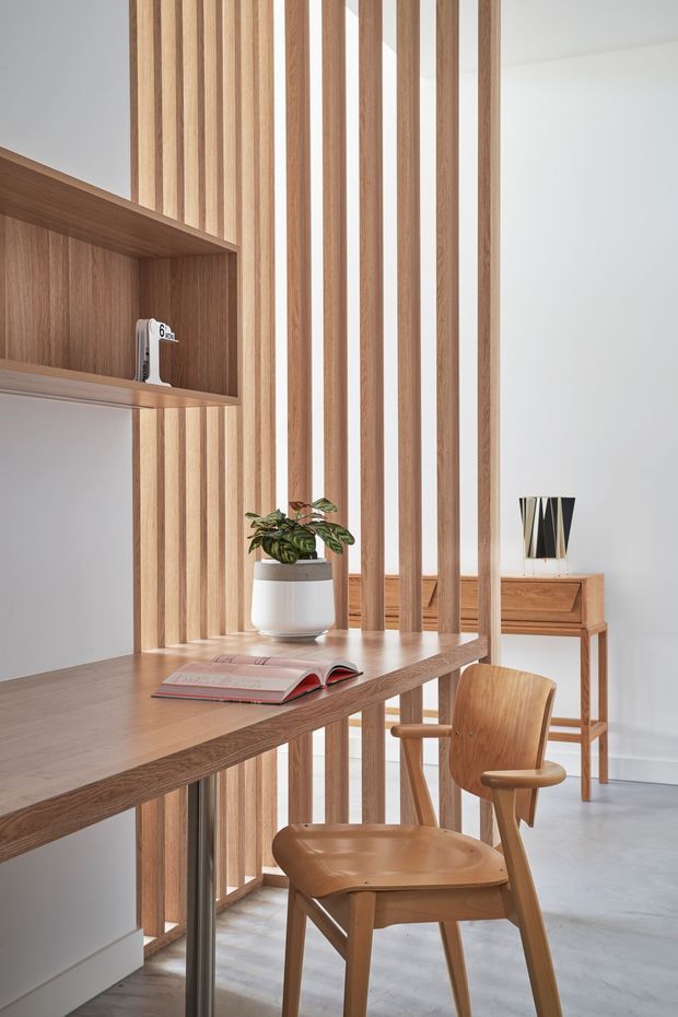 A refined home workspace with a built-in timber desk extending from wall shelving, framed by a vertical timber slat divider. A wooden chair, potted plant, open book, and small console table with decorative lamp complete the bright, minimalist setting. Pale walls and a light concrete-like floor contrast with the warm natural wood joinery.