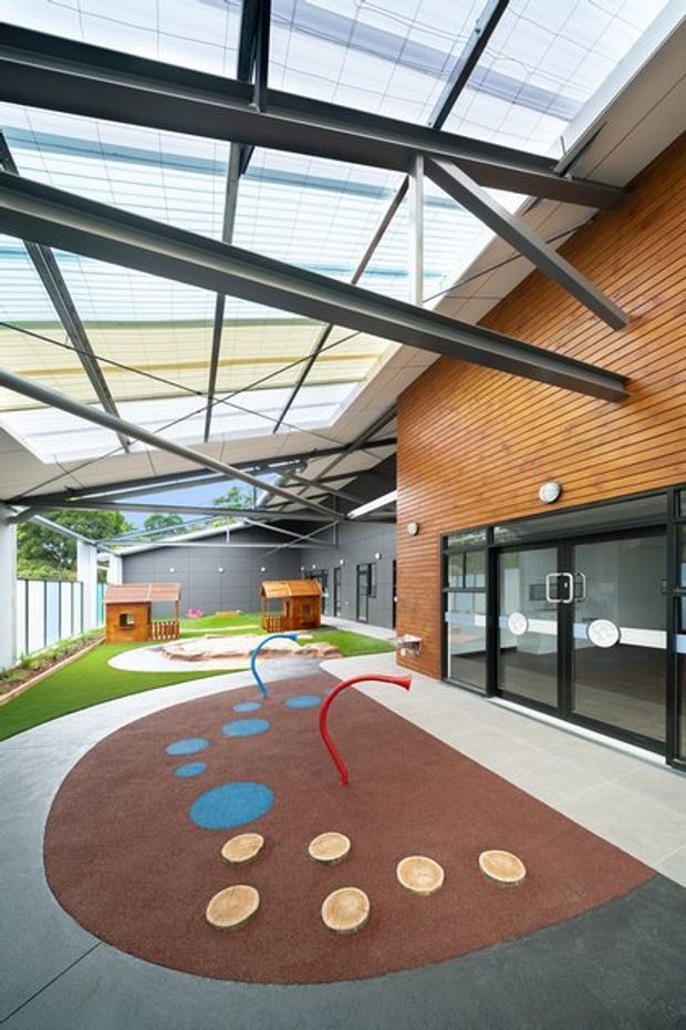 A covered outdoor play courtyard attached to a contemporary building, featuring a translucent roof canopy supported by exposed dark steel beams, a warm timber-clad wall, black-framed glazed doors, and colourful rubber play surfacing with stepping discs and curved play elements. Artificial grass, play structures, and a landscaped outdoor area are visible beyond.