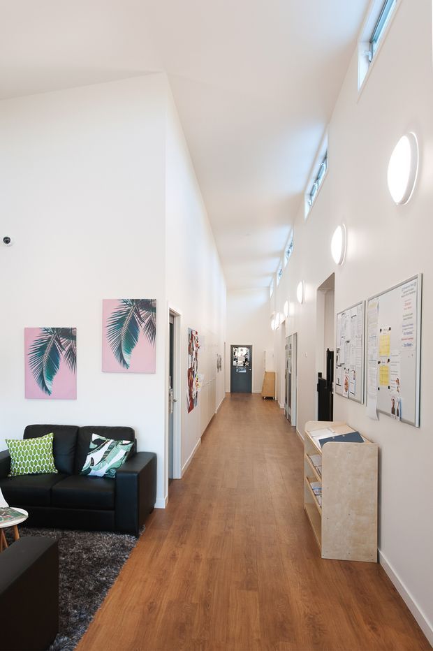 Bright, contemporary interior hallway with a long timber-look floor, white walls, high ceiling and clerestory windows. A black sofa and tropical artwork create a small waiting or lounge area on the left, while noticeboards, doors and built-in storage line the corridor. Round wall lights provide repeated illumination along the right wall.