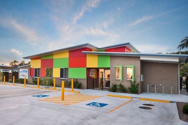 A bright, low-rise commercial building with a playful multicoloured panel facade, dark brick walls, a shallow sloping roof and sheltered entrance. The foreground contains a concrete parking and accessible drop-off area with yellow bollards and marked accessible spaces. Palm trees and clear sky are visible in the background.