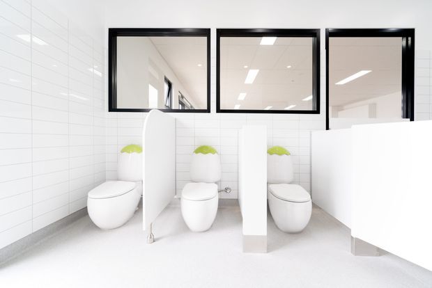 Bright, minimalist commercial bathroom with three white wall-mounted toilets separated by curved white privacy partitions. White horizontal wall tiles, pale flooring, black-framed interior windows, and bright recessed ceiling lighting create a clean, high-key public washroom.