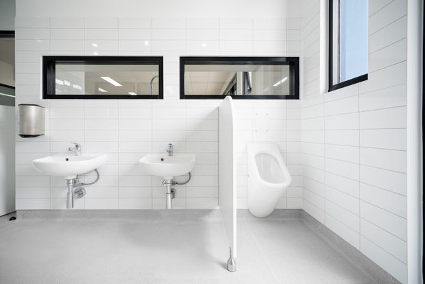 Bright, minimalist commercial bathroom with white rectangular wall tiles, two wall-mounted basins, exposed chrome plumbing, a wall-mounted urinal, and a white privacy partition. The space has a grey seamless floor, black-framed horizontal windows, and clean contemporary fittings.