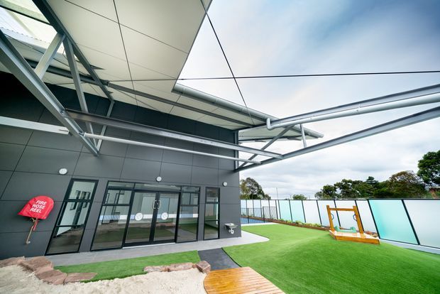 Modern outdoor recreational courtyard beside a dark-clad building, featuring extensive steel framing and exposed structural beams, a translucent roof or canopy, full-height glazed doors, artificial turf, a colorful boundary screen, and a timber walkway. The space appears to be a covered outdoor play or activity area.