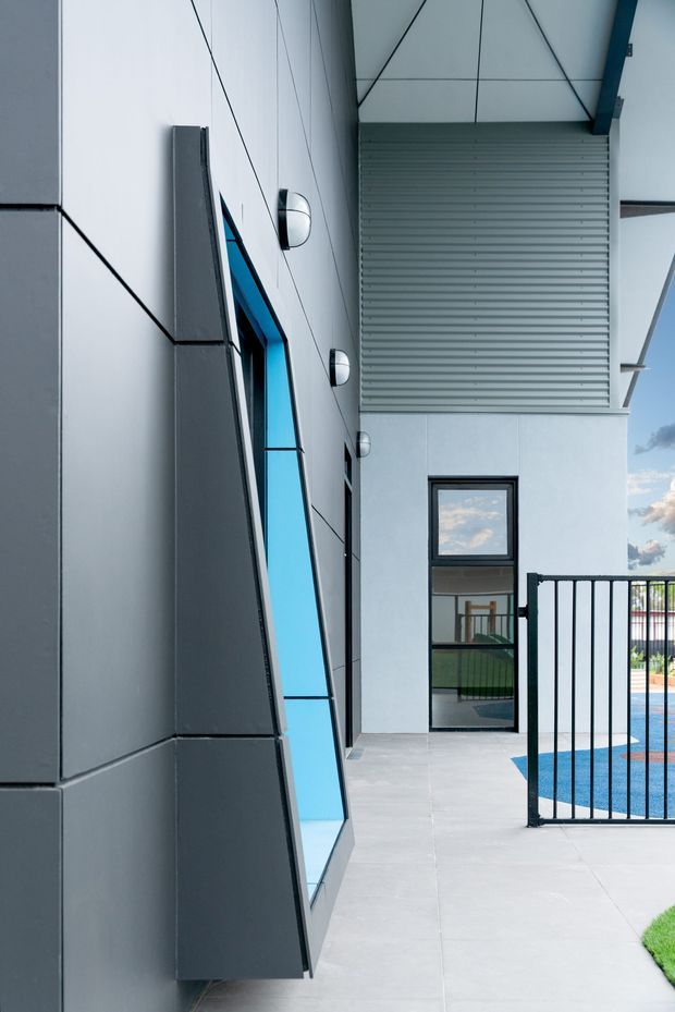 A modern exterior courtyard or poolside passage featuring dark grey geometric wall cladding, a striking angled blue-tinted window, smooth pale concrete paving, black metal balustrading, and a partially visible bright blue swimming pool. The building has clean contemporary lines, exposed outdoor wall lights, and a projecting louvered or ribbed upper facade element.