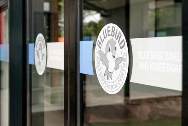 Close-up exterior view of glazed entry doors featuring Bluebird Early and Kindergarten branding, including a circular bird logo and frosted horizontal signage bands. Reflections and a subdued building entrance are visible in the glass.