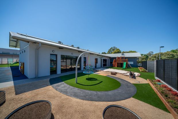 Bright exterior view of a contemporary single-storey building surrounding a landscaped play courtyard. The white rendered building has a low-pitched roof, large glazed doors and windows, and a covered walkway. The courtyard includes artificial turf, rubber play surfacing, a curved metal play structure, swings, a slide, sand areas, raised garden beds, and dark privacy fencing.
