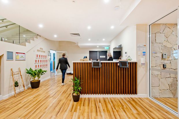 Bright contemporary reception lobby with a timber-look floor and a prominent reception desk featuring vertical dark timber slats and a light stone countertop. The space includes recessed ceiling lighting, a glass-partitioned stairway on the left, indoor plants, wall-mounted artwork and notices, and a large stone-clad feature wall and glazed doorway on the right.