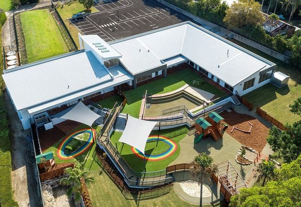 Aerial view of a large single-storey institutional or educational building with several white metal-clad wings arranged around a central landscaped courtyard. The grounds include a curved tiered seating area, covered outdoor play spaces, colourful circular play markings, timber play equipment, sand or mulch areas, fenced recreation zones, and an adjacent marked parking lot.