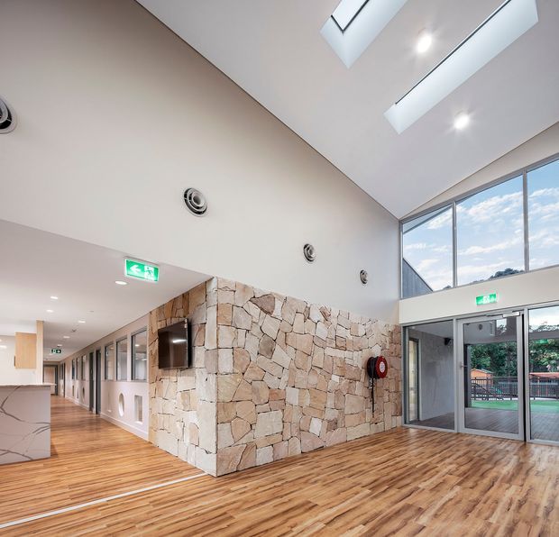 Bright contemporary institutional interior lobby with a tall angled ceiling, skylights, clerestory glazing, and full-height windows. A prominent irregular natural-stone feature wall incorporates a wall-mounted screen, while timber-look flooring runs through the open circulation space. Glazed entry doors, visible offices or rooms along the corridor, recessed lighting, ventilation grilles, and illuminated exit signs complete the setting.