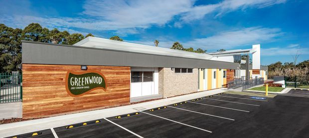 Single-storey contemporary commercial building viewed from the front, with a low flat roof, dark grey rendered facade, horizontal timber-clad feature wall bearing a Greenwood sign, extensive glazing, and a newly marked asphalt parking area. The property is set against trees and a bright sky.