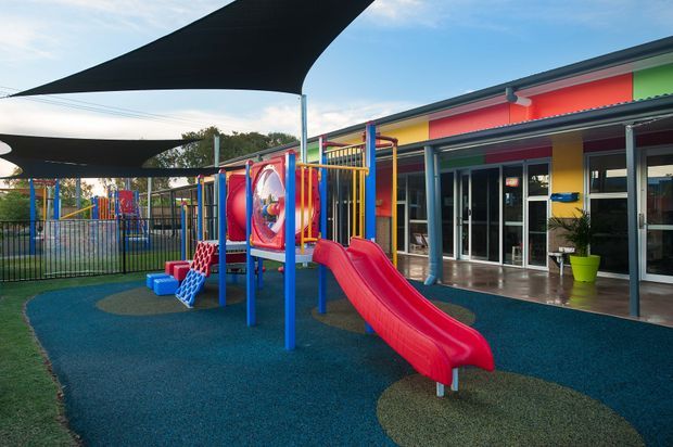 A colourful outdoor children's playground beside a single-storey building, featuring a bright red slide, climbing structures, play tunnels, safety fencing, and shaded areas beneath large tensile shade sails. The building has vivid multicoloured facade panels, glazed doors and windows, and a covered veranda.