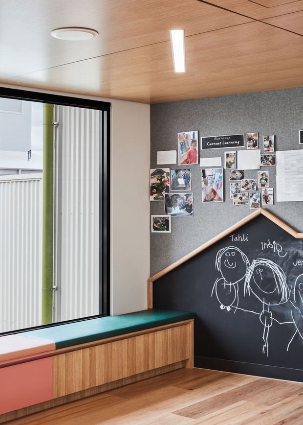 A bright, compact children's learning or activity room with a full-height black-framed window, timber-lined ceiling, warm timber flooring, and a built-in window seat with colourful upholstered sections and timber cabinetry below. One wall combines grey felt pinboards displaying children's artwork and notices with a large house-shaped chalkboard featuring hand-drawn figures. The room has a playful, contemporary educational character with warm natural materials.
