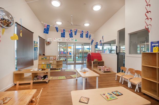 Bright, spacious early-learning classroom or childcare room with timber-look flooring, low children's tables and chairs, open shelving, educational toys, a play mat, and large glazed doors and windows. Australian-themed bunting and children's artwork decorate the room. The space has a clean, practical, welcoming fit-out with abundant daylight and recessed ceiling lights.