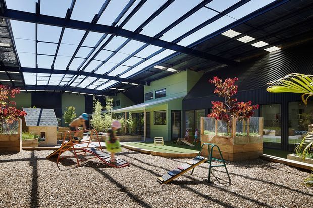 A contemporary enclosed outdoor courtyard or play area with a gravel surface, timber planter boxes, children’s play equipment, and lush planting. The space is surrounded by dark corrugated-clad and green-painted building volumes, with extensive overhead steel framing and translucent roof panels creating a covered, naturally lit setting.