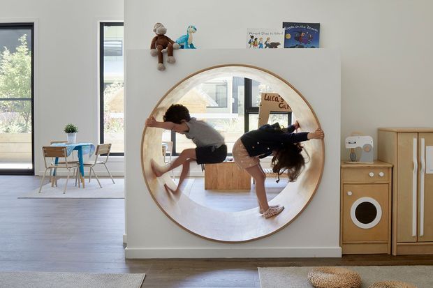 Bright, playful children's playroom with a large round wall mirror, a small children's table and chairs, toys and books on a wall ledge, and built-in wooden toy-kitchen cabinetry. Two children are reflected in the mirror, with additional light-filled living space visible beyond.