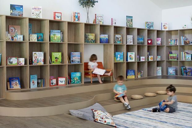 A bright, contemporary children's reading and play area with extensive built-in timber-look shelving filled with books, stepped tiered seating, and a small integrated bench. Children are reading and sitting on cushions and a soft rug in the foreground. The space has white walls, warm wood-toned finishes, and a playful, welcoming library-like atmosphere.