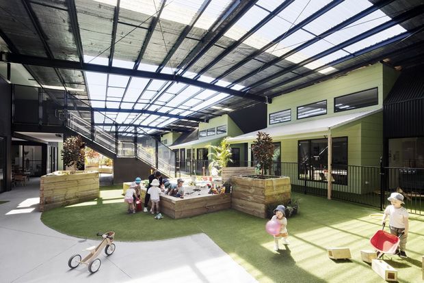 A bright covered outdoor courtyard or play area enclosed by contemporary low-rise buildings. The space features a large translucent and open steel roof structure, concrete paths, artificial grass, raised timber planter boxes, children’s play equipment, and planted areas. The surrounding building has dark and lime-green horizontal cladding, extensive glazing, and metal railings, creating an institutional or childcare setting.