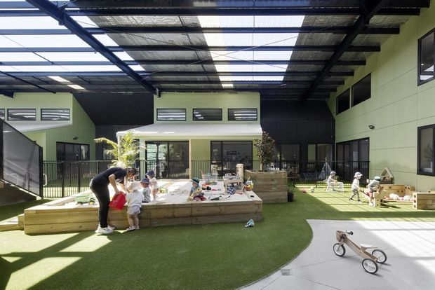A large, covered outdoor play area within a contemporary childcare or educational facility. The space has artificial turf, raised timber planter and sand-play structures, children's play equipment, black metal fencing, pale green and black wall cladding, and extensive translucent roof panels that provide bright natural light. Children and an adult are using the play area.