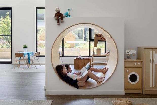 Bright contemporary children's playroom with a large circular timber-framed wall mirror, built-in or fixed wooden toy cabinetry, and a recessed reading nook visible in the mirror reflection. Two children are relaxing with a book in the nook. The room has pale walls, light timber flooring, large black-framed windows, and a small children's table and chairs in the adjoining area.