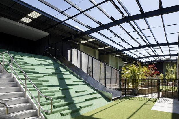 A bright, contemporary semi-outdoor architectural space with a large green artificial-turf stepped ramp or amphitheatre seating area beside a conventional stair. Black steel framing supports a transparent roof canopy, with metal handrails, mesh balustrades, and planted landscaping in a lower courtyard.