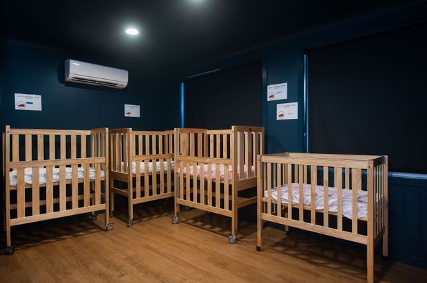 A dark navy-painted nursery or childcare room displaying several wooden baby cribs with casters, arranged across a warm timber-look floor. The room has roller blinds, recessed ceiling lighting, wall-mounted air conditioning, and small informational signs.