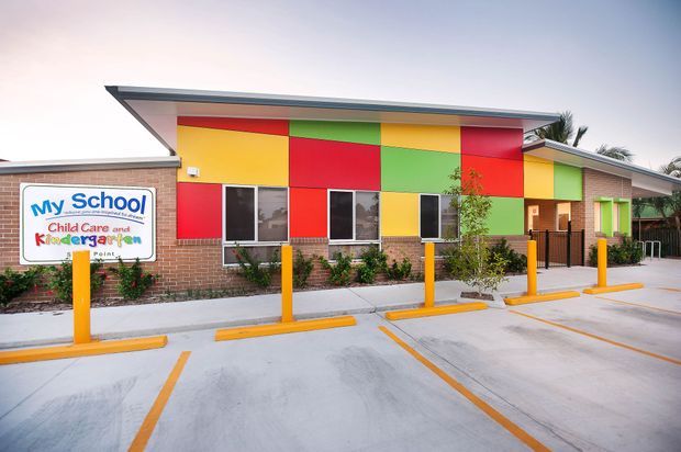 Exterior view of a colourful childcare and kindergarten building, with a low-slung roof, exposed brick lower walls, bright red, yellow, green and orange facade panels, windows, a fenced entrance, and a marked concrete parking area with yellow bollards.