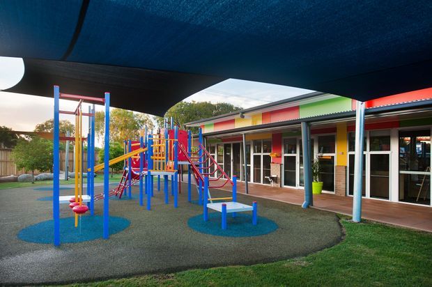 Colourful outdoor playground beside a low-rise building, with bright blue, red, yellow and green play equipment, rubber safety surfacing, a broad shade canopy, and a covered veranda with glazed doors and vivid facade panels.