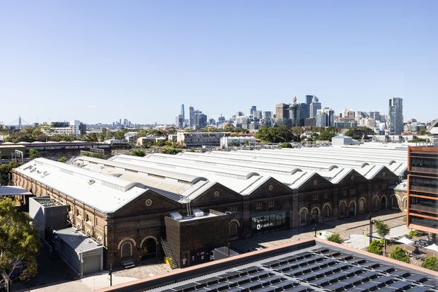 Wide elevated exterior view of a large historic brick industrial or market building with repeated arched façades, ornate circular details, and multiple sawtooth roof sections. Modern city high-rises are visible in the background, with rooftop solar panels in the foreground and an adjacent contemporary building at right.