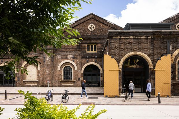 Exterior view of a historic dark-brick institutional or commercial building with prominent arched openings, ornate brick detailing, a central gable marked with the number 10, and large mustard-yellow timber doors. The streetscape includes paving, bollards, bicycles, pedestrians, and landscaped greenery. The image is strongly focused on the preserved brick facade and entry architecture.