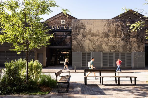 Exterior view of a contemporary adaptive-reuse style building in a landscaped urban plaza. The facade combines dark brick gabled warehouse-like volumes, horizontal metal louvres, a large weathered mural or graphic wall panel, and a recessed pedestrian entrance. Benches, paving, planting, and people occupy the foreground.