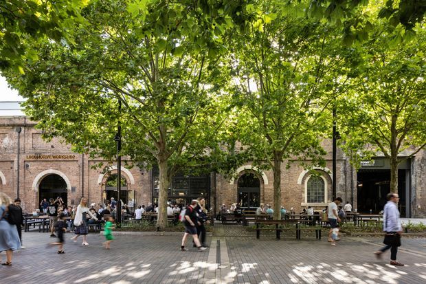 Street-level view of a leafy urban pedestrian precinct in front of a restored brick masonry building. Several arched openings, tall arched windows, dark shopfronts, and outdoor café seating line the facade, with people walking beneath a dense canopy of mature trees. The scene has a lively adaptive-reuse or commercial-dining character.