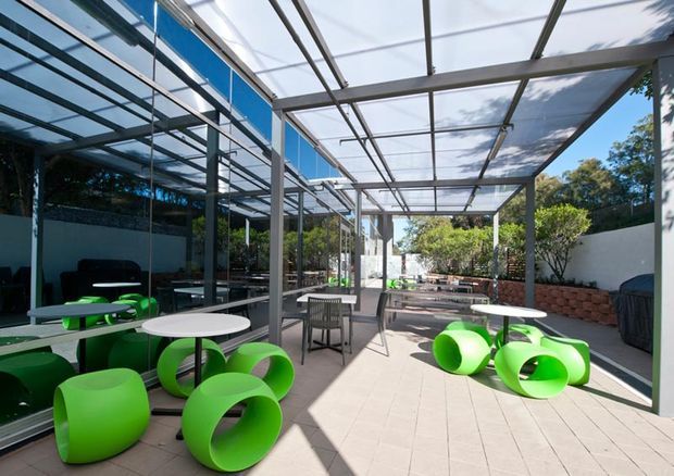 A bright contemporary outdoor terrace or courtyard beside a highly glazed building, sheltered by a transparent pergola with a metal frame. The paved space contains vivid lime-green sculptural seating, white round tables, and a central dining table with chairs. White rendered walls, brick planter edging, greenery, and strong sunlight create a modern hospitality-style outdoor setting.