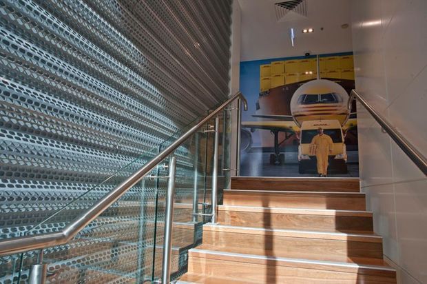 A modern commercial stairway with warm timber stair treads, stainless-steel handrails, and clear glass balustrades. A patterned perforated metal screen forms the left wall, while a large aviation-themed mural is visible at the upper landing. The space is brightly lit with a clean, utilitarian interior fit-out.