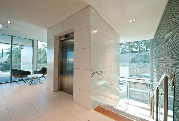 Bright contemporary interior circulation space with a stainless-steel elevator set into a large pale tiled volume. Full-height glazing overlooks greenery, while a stair with stainless-steel handrails descends beside a dramatic ribbed feature wall and timber stair treads. Minimal lounge seating is visible in the adjoining glazed area.
